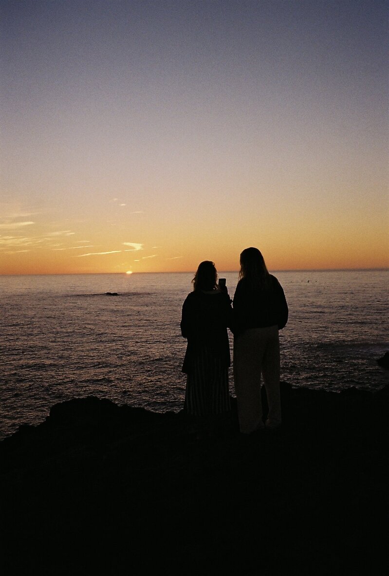 Twee vrouwen bij zonsondergang aan zee, een maakt een foto met haar telefoon